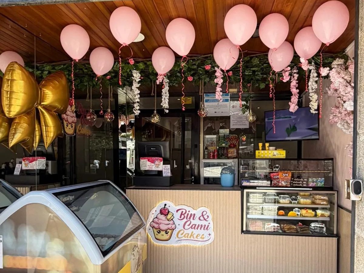 El mostrador de Bin & Cami Cakes visto desde la calle: globos rosas, el cartel iluminado con el cupcake, la vitrina de dulces y el arcón de los helados.