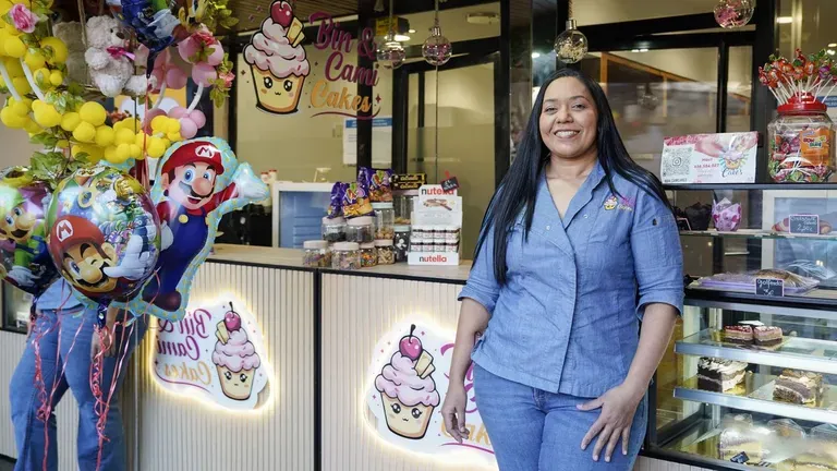 Una de las pasteleras, sonriendo junto al mostrador de Bin & Cami Cakes, con el rótulo iluminado de la marca detrás y la vitrina de dulces al fondo.