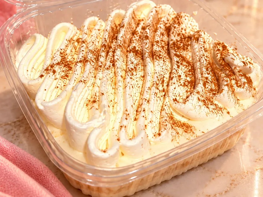 Tarta tres leches en su envase, cubierta de nata montada a la manga y espolvoreada de canela